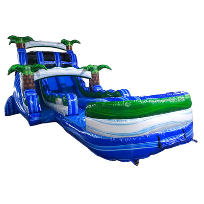 Large tropical-themed inflatable water slide with blue base and green accents, featuring dual lanes, palm tree decorations, and an extended splash pool with wave design.