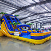 Angled view of 18ft Lava Falls Center Climb water slide featuring blue sliding lanes with yellow borders, inflatable palm trees, central climbing structure, and a spacious splash pool, displayed in a large indoor facility with metal roofing and lighting visible.