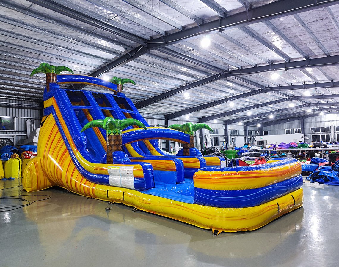 Angled view of 18ft Lava Falls Center Climb water slide featuring blue sliding lanes with yellow borders, inflatable palm trees, central climbing structure, and a spacious splash pool, displayed in a large indoor facility with metal roofing and lighting visible.