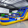 Side angle view of the 18ft Lava Falls inflatable water slide, showcasing its blue sliding lanes, yellow accents, palm tree decorations, and large splash pool area, set up in an indoor warehouse with metal ceiling and other inflatables in the background.