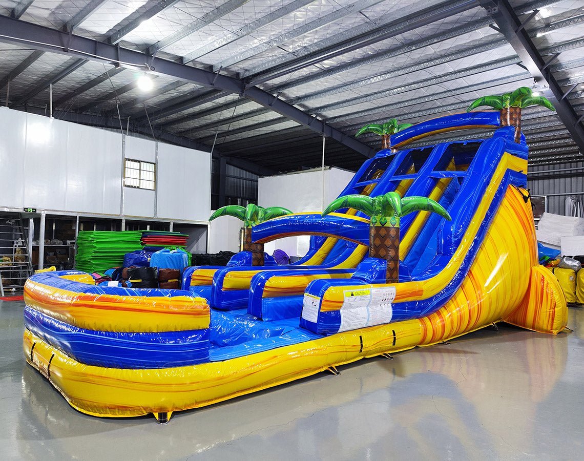 Side angle view of the 18ft Lava Falls inflatable water slide, showcasing its blue sliding lanes, yellow accents, palm tree decorations, and large splash pool area, set up in an indoor warehouse with metal ceiling and other inflatables in the background.