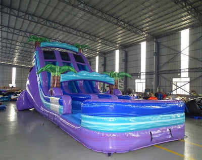 Side view of 18ft Purple Plunge Center Climb water slide showing vibrant purple, blue, and turquoise colors, inflatable palm trees, and a large splash pool area, set up in an indoor warehouse with metal roofing and other inflatables visible.