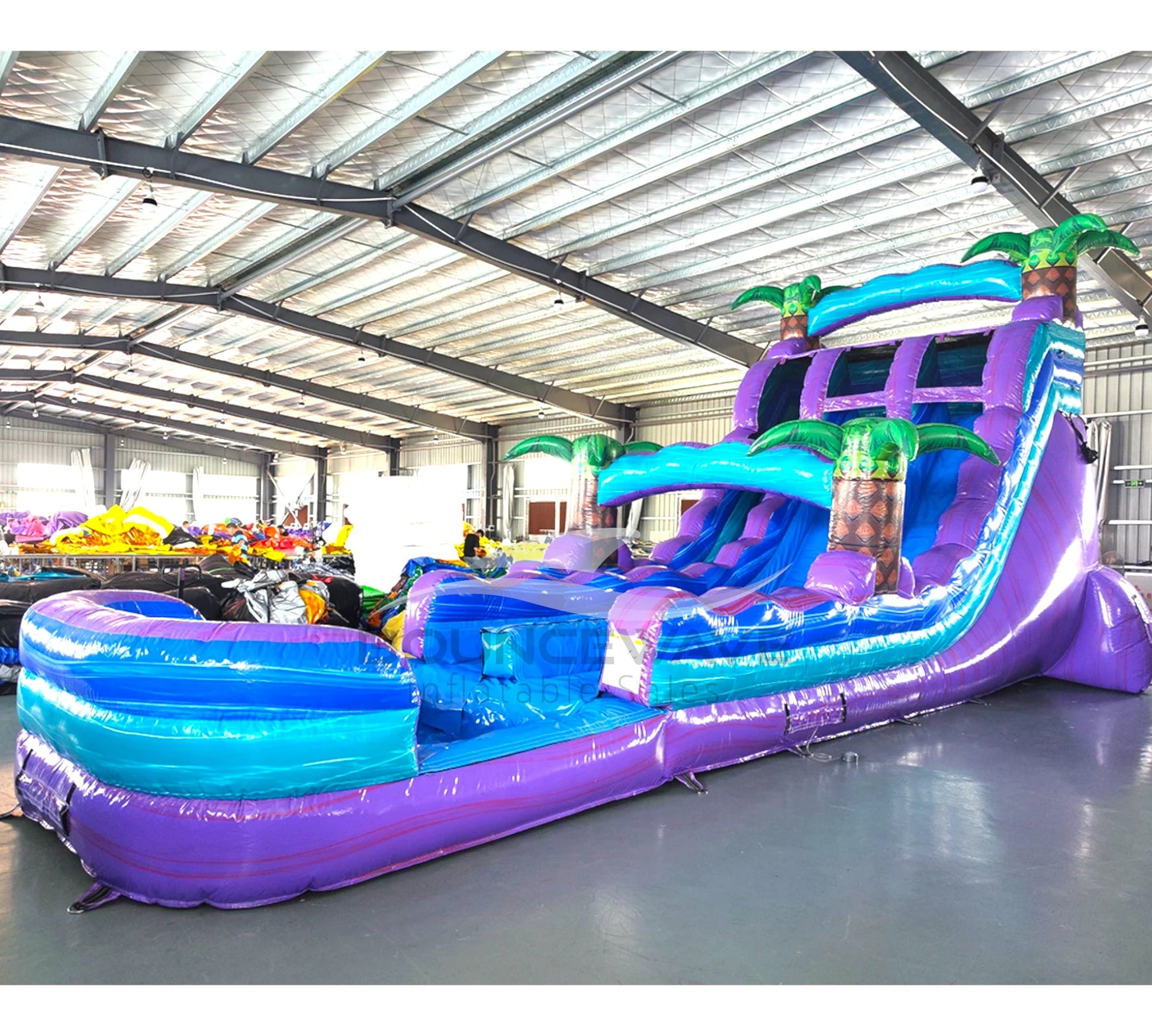 Large tropical-themed inflatable water slide with purple and blue colors, featuring dual lanes, palm tree decorations, and an extended splash pool in indoor warehouse setting.