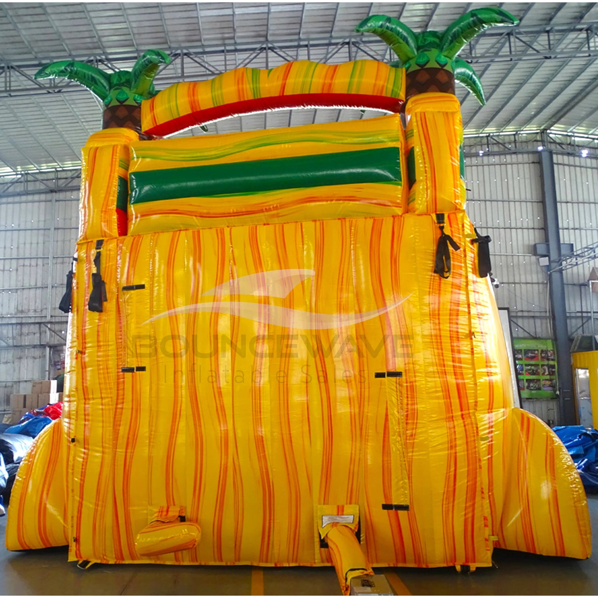 Large tropical-themed inflatable water slide with yellow base, featuring dual lanes, palm tree decorations, and an extended splash pool in indoor warehouse setting.