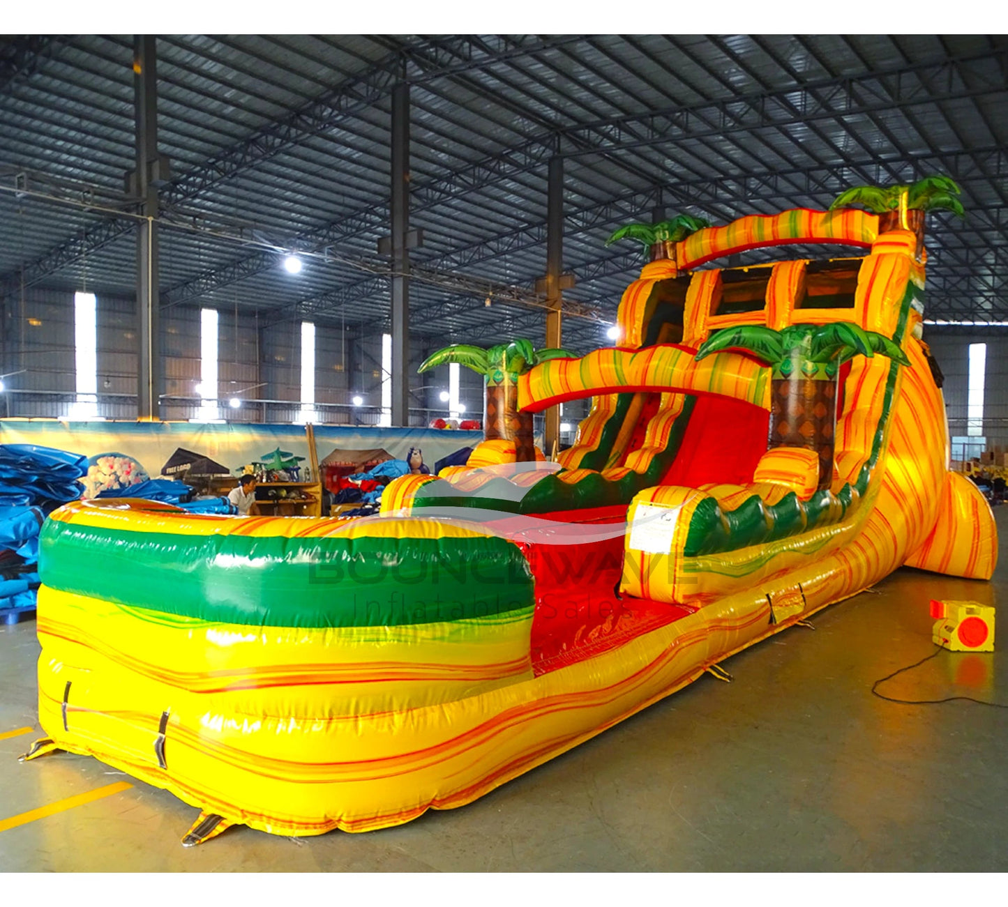 Angled view of Rasta Splash hybrid water slide in indoor warehouse, showcasing its height, dual lanes, tropical theme with palm trees, and extended yellow and green splash pool.