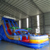 Large inflatable water slide with tropical theme, featuring blue sliding lanes, palm tree decorations, and a colorful splash pool area, set up in an indoor warehouse with metal roofing visible.