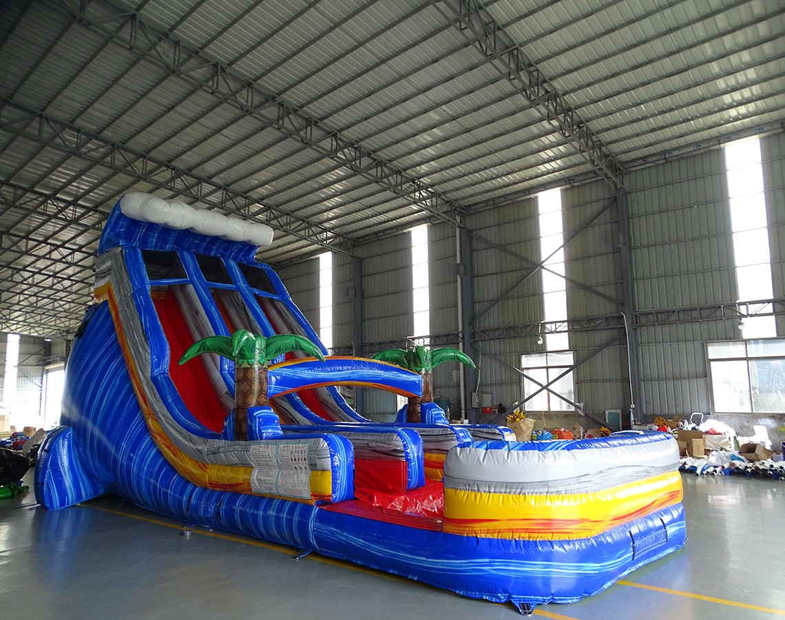 Large inflatable water slide with tropical theme, featuring blue sliding lanes, palm tree decorations, and a colorful splash pool area, set up in an indoor warehouse with metal roofing visible.