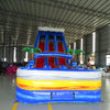 Front view of the 18ft Rip Curl inflatable water slide, showcasing its blue sliding lanes, palm tree decorations, and colorful splash pool, set up in an indoor warehouse with other inflatables and equipment visible in the background.