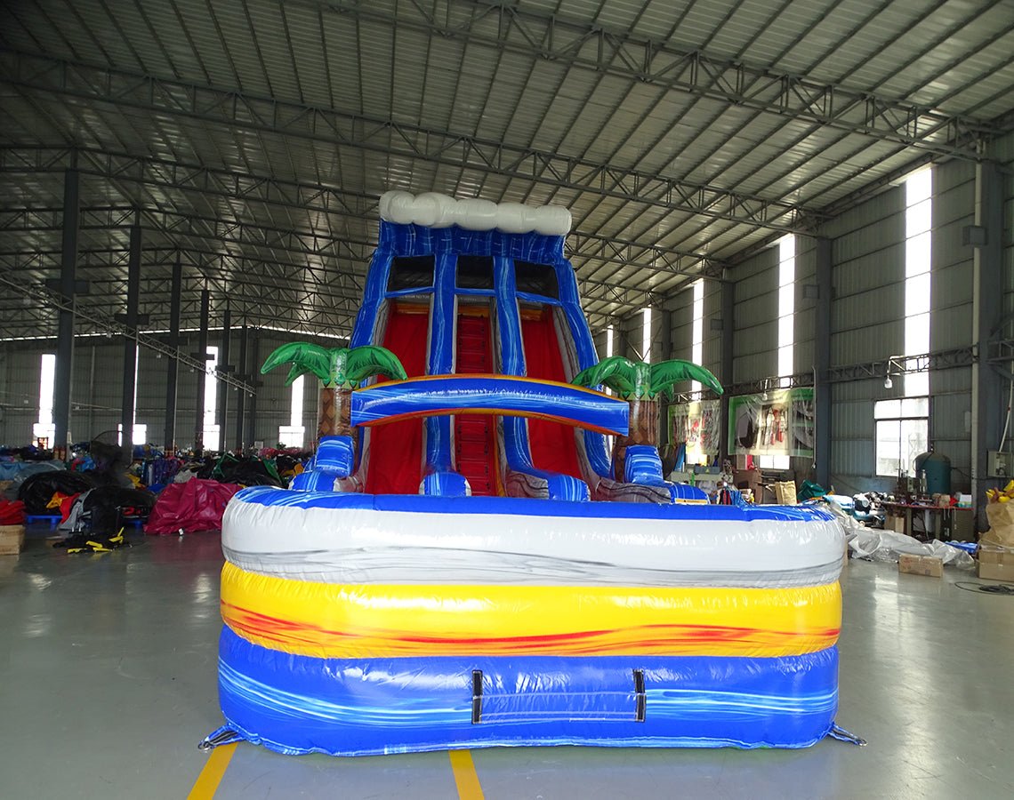 Front view of the 18ft Rip Curl inflatable water slide, showcasing its blue sliding lanes, palm tree decorations, and colorful splash pool, set up in an indoor warehouse with other inflatables and equipment visible in the background.