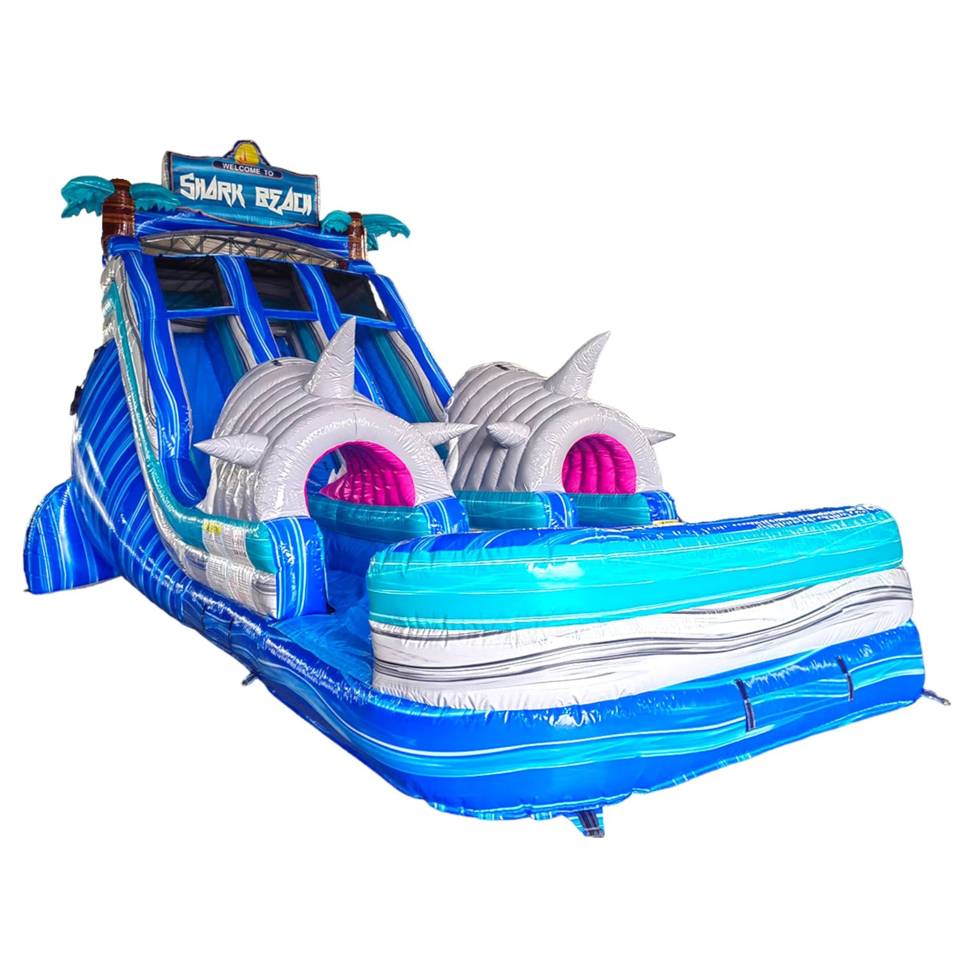 Isolated view of the 18ft Shark Beach inflatable water slide, showcasing its blue sliding lanes, shark head obstacles, palm tree decorations, and "Welcome to Shark Beach" sign on a white background.