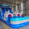 Side view of 18ft Shark Beach Center Climb water slide showing blue sliding lanes, inflatable shark heads, palm tree decorations, and a large splash pool area with blue and white wave patterns, displayed in an indoor facility.