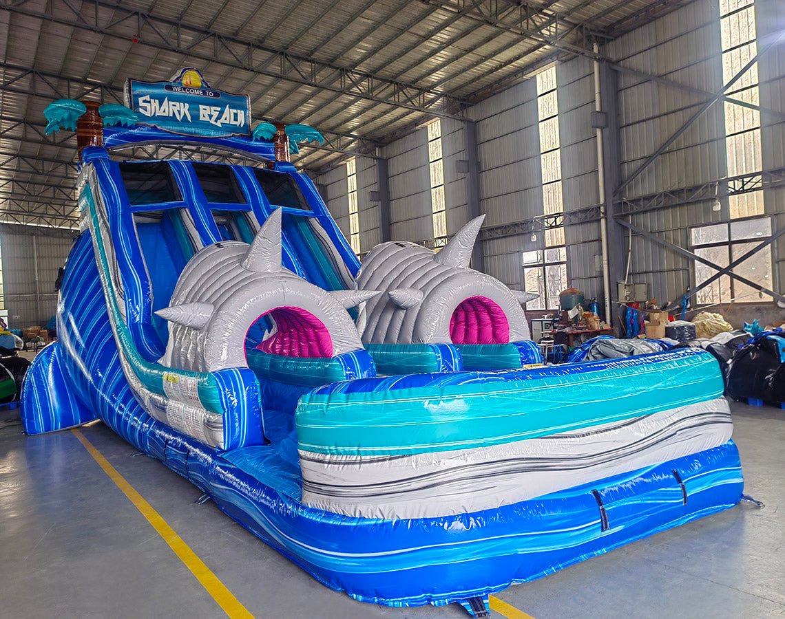 Side view of 18ft Shark Beach Center Climb water slide showing blue sliding lanes, inflatable shark heads, palm tree decorations, and a large splash pool area with blue and white wave patterns, displayed in an indoor facility.