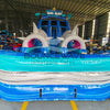 Front view of 18ft Shark Beach Center Climb water slide featuring blue sliding lanes, inflatable shark heads with pink mouths, palm tree decorations, and a large splash pool with "BOUNCEWAVE Inflatable Sales" branding, set up in an indoor warehouse surrounded by other inflatables.