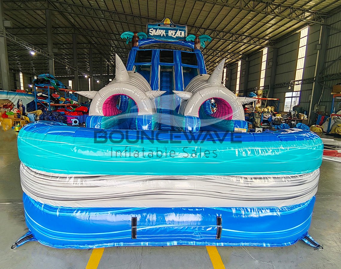 Front view of 18ft Shark Beach Center Climb water slide featuring blue sliding lanes, inflatable shark heads with pink mouths, palm tree decorations, and a large splash pool with "BOUNCEWAVE Inflatable Sales" branding, set up in an indoor warehouse surrounded by other inflatables.