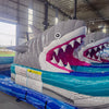 Large inflatable water slide with shark theme, featuring blue sliding lanes, two inflatable shark heads with open mouths, palm tree decorations, and a "Welcome to Shark Beach" sign at the top. The slide has a spacious splash pool area and is set up in an indoor warehouse.