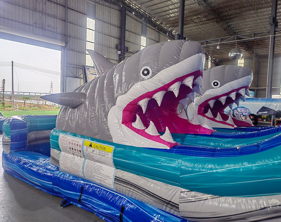 Large inflatable water slide with shark theme, featuring blue sliding lanes, two inflatable shark heads with open mouths, palm tree decorations, and a "Welcome to Shark Beach" sign at the top. The slide has a spacious splash pool area and is set up in an indoor warehouse.