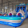 Large inflatable water slide with shark theme, featuring blue sliding lanes, inflatable shark heads with pink mouths, palm tree decorations, and a "Welcome to Shark Beach" sign, set up in an indoor warehouse.