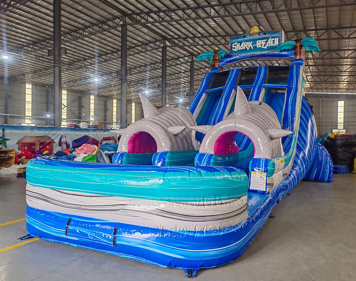 Large inflatable water slide with shark theme, featuring blue sliding lanes, inflatable shark heads with pink mouths, palm tree decorations, and a "Welcome to Shark Beach" sign, set up in an indoor warehouse.