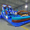 Angled view of the 18ft South Beach inflatable water slide, showcasing its blue sliding lanes, palm tree decorations, "South Beach" surfboard sign, and large splash pool area, set up in an indoor warehouse with other equipment visible.