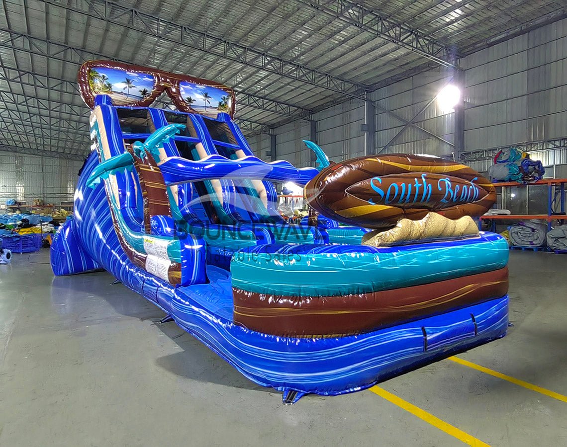 Angled view of the 18ft South Beach inflatable water slide, showcasing its blue sliding lanes, palm tree decorations, "South Beach" surfboard sign, and large splash pool area, set up in an indoor warehouse with other equipment visible.