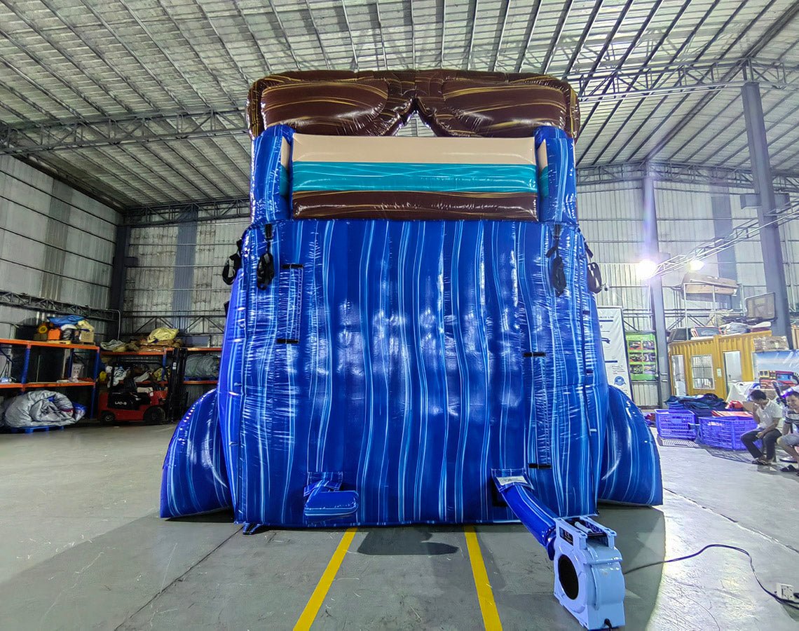 Large inflatable water slide with tropical theme, featuring blue sliding lanes, palm tree decorations, a "South Beach" surfboard sign, and a colorful splash pool area. The slide has a beach scene backdrop and is set up in an indoor warehouse with metal roofing visible.