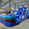 Large inflatable water slide with tropical theme, featuring blue sliding lanes, palm tree decorations, and a "South Beach" surfboard sign, set up in an indoor warehouse with metal roofing and other inflatables visible.