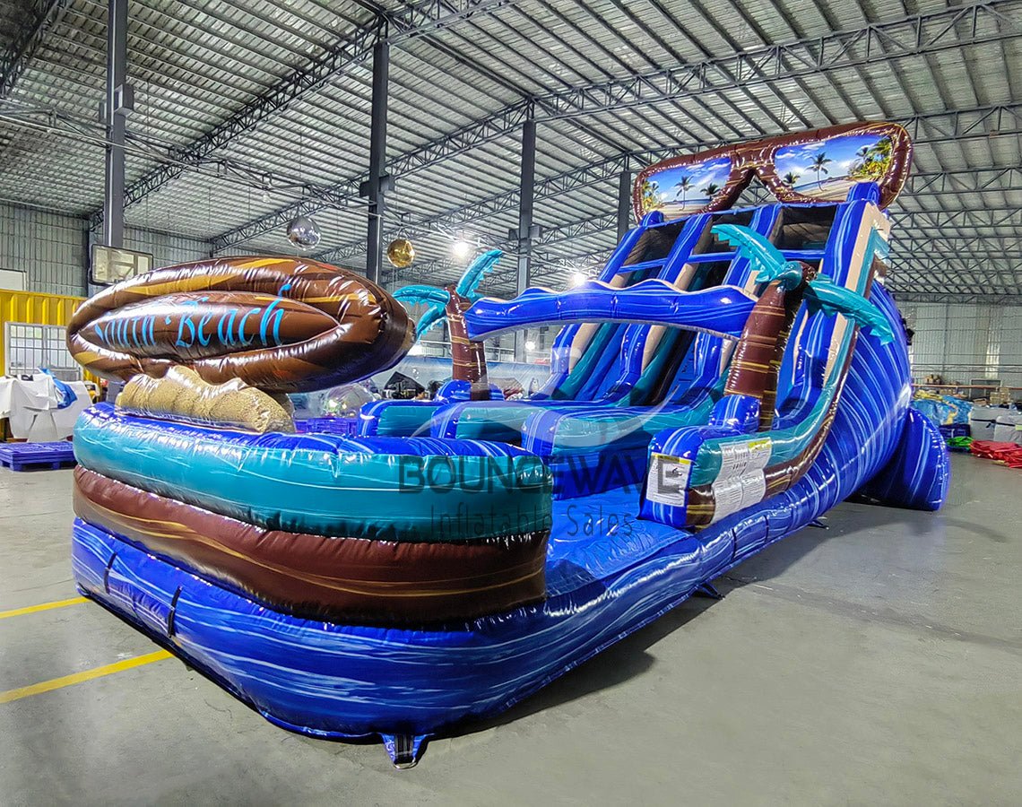 Large inflatable water slide with tropical theme, featuring blue sliding lanes, palm tree decorations, and a "South Beach" surfboard sign, set up in an indoor warehouse with metal roofing and other inflatables visible.