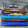 Front view of 18ft South Beach Center Climb water slide featuring blue sliding lanes, inflatable palm trees, tropical scene backdrop, and a large "South Beach" surfboard sign, displayed in an indoor facility with other inflatables and workers visible in the background.