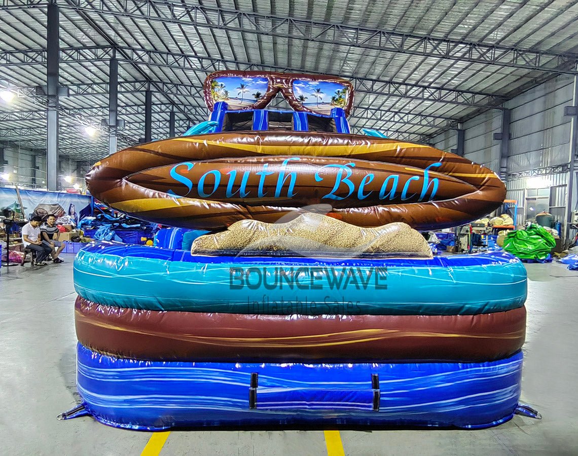 Front view of 18ft South Beach Center Climb water slide featuring blue sliding lanes, inflatable palm trees, tropical scene backdrop, and a large "South Beach" surfboard sign, displayed in an indoor facility with other inflatables and workers visible in the background.