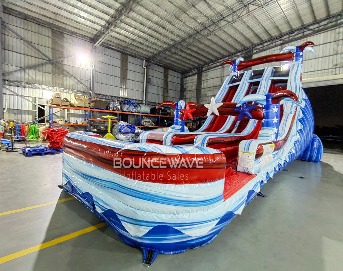 Angled view of the 18ft Star Spangled Splash inflatable water slide, showcasing its American flag-inspired design with red, white, and blue colors, star decorations, three sliding lanes, and a large splash pool area with "BOUNCEWAVE Inflatable Sales" branding.