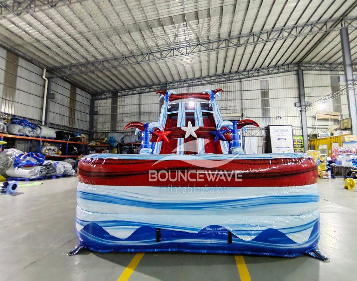 Front view of 18ft Star Spangled Splash Center Climb water slide featuring patriotic color scheme, star decorations, three sliding lanes, and a spacious splash pool with red, white, and blue wave patterns, set up in an indoor warehouse surrounded by other inflatables and equipment.