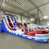 Large inflatable water slide with American flag theme, featuring red, white, and blue colors, star decorations, and three sliding lanes, set up in an indoor warehouse with metal roofing and other inflatables visible.