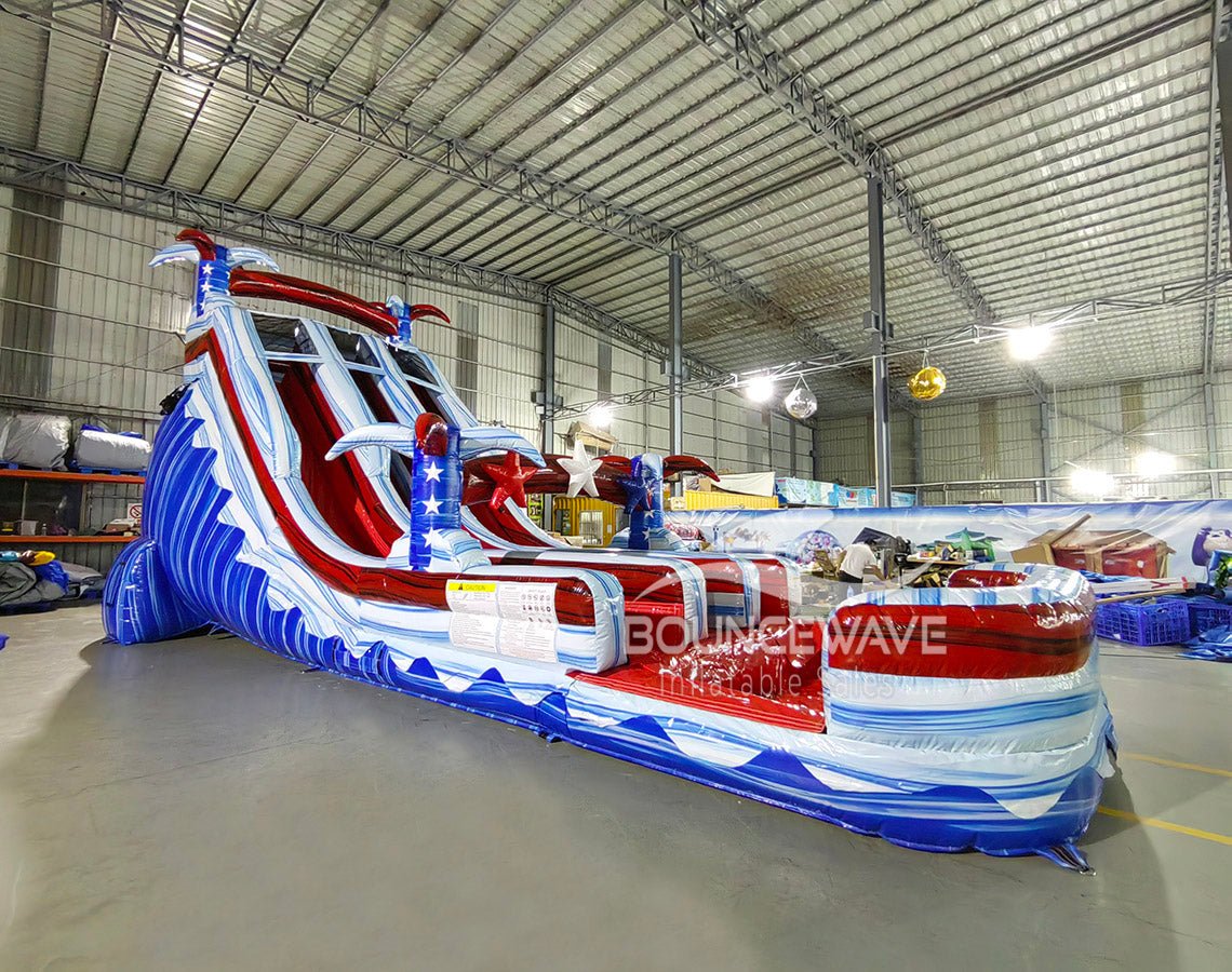 Large inflatable water slide with American flag theme, featuring red, white, and blue colors, star decorations, and three sliding lanes, set up in an indoor warehouse with metal roofing and other inflatables visible.