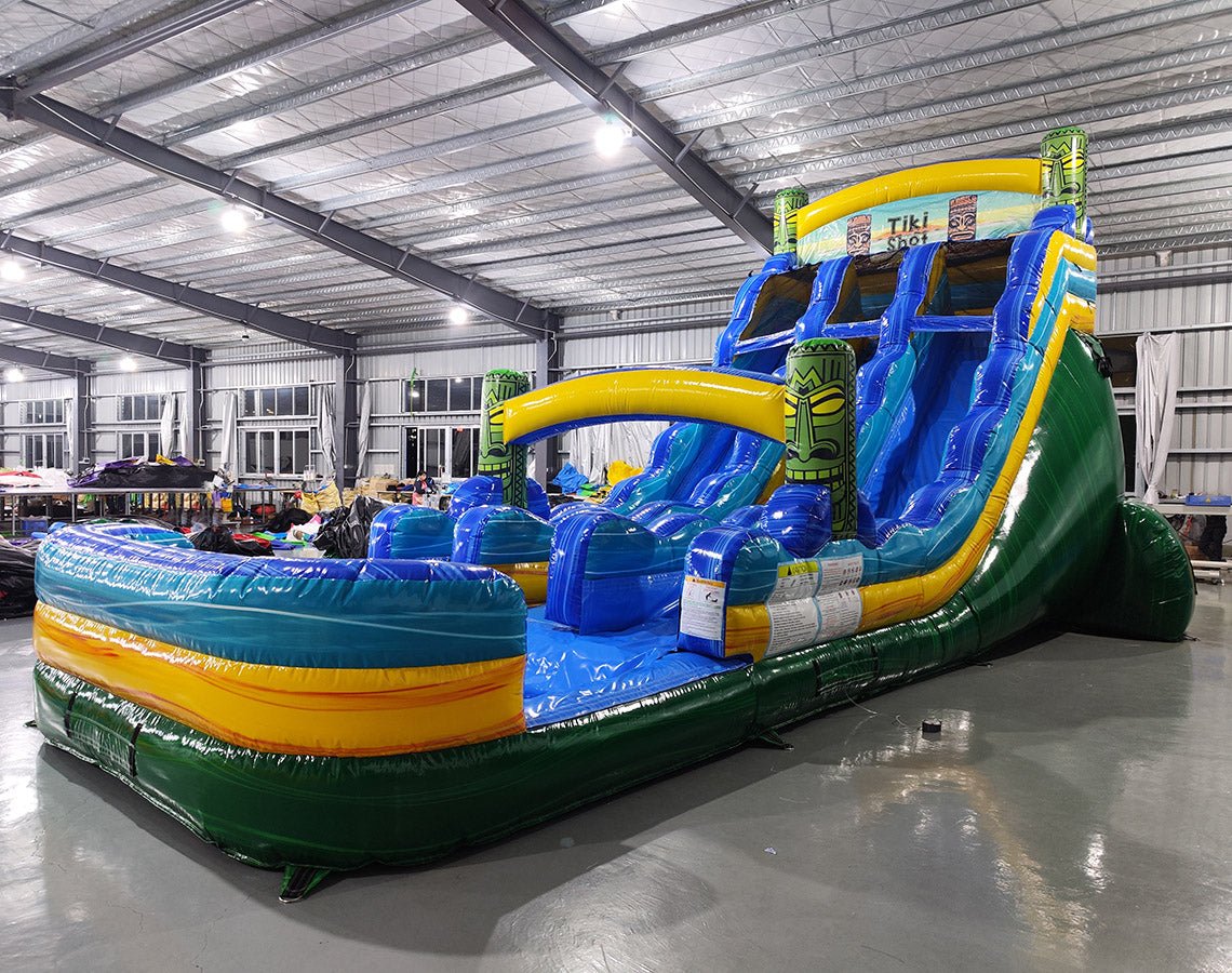 Side view of 18ft Tiki Shot Center Climb water slide showing vibrant blue, yellow, and green colors, tiki decorations, and a large splash pool area, displayed in a spacious indoor facility with metal roofing.