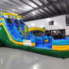 Large inflatable water slide with tropical theme, featuring blue sliding lanes, yellow and green accents, tiki decorations, and a spacious splash pool area. The slide is set up in an indoor warehouse with metal roofing and other inflatables visible.