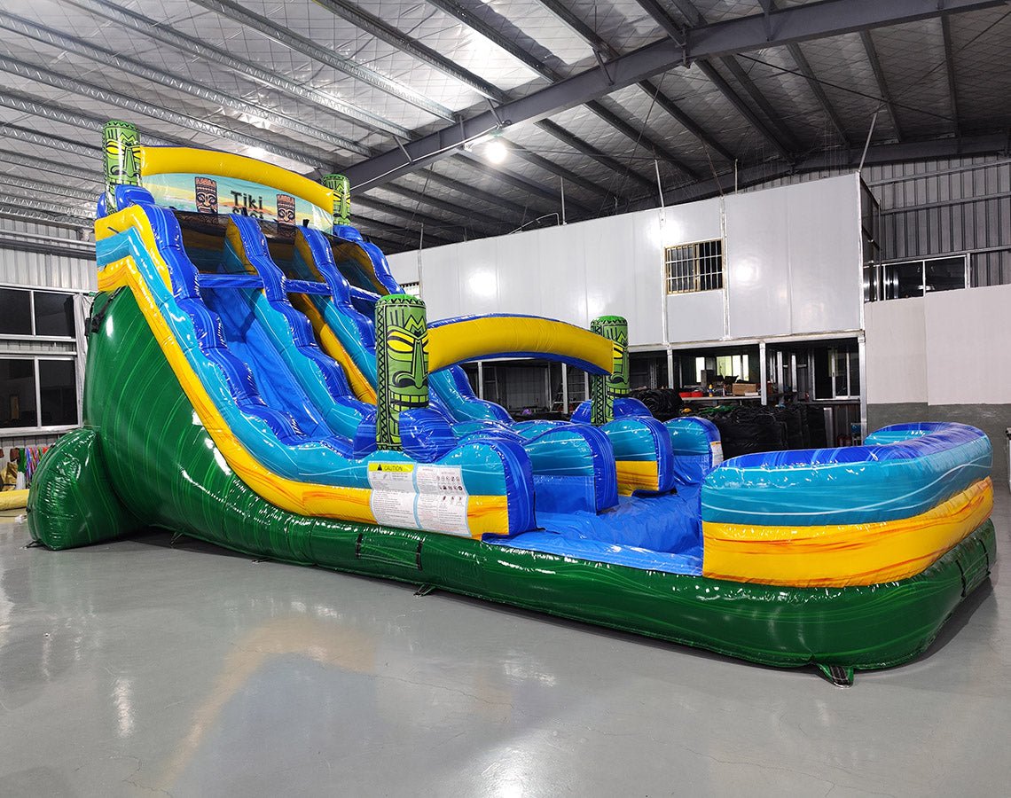 Large inflatable water slide with tropical theme, featuring blue sliding lanes, yellow and green accents, tiki decorations, and a spacious splash pool area. The slide is set up in an indoor warehouse with metal roofing and other inflatables visible.