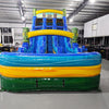 Front view of the 18ft Tiki Shot inflatable water slide, showcasing its blue sliding lanes, tiki decorations, yellow accents, and large splash pool area. The slide is set up in an indoor warehouse with other inflatables and equipment visible in the background.