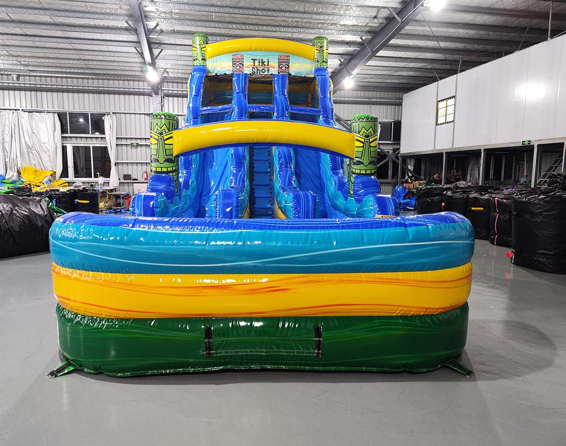 Front view of the 18ft Tiki Shot inflatable water slide, showcasing its blue sliding lanes, tiki decorations, yellow accents, and large splash pool area. The slide is set up in an indoor warehouse with other inflatables and equipment visible in the background.