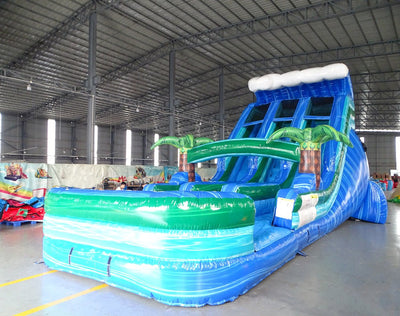 Side angle view of 18ft Trop Wave Center Climb water slide showing blue sliding lanes, inflatable palm trees, and a large splash pool area. The slide is set up in an indoor facility with other inflatables visible in the background.