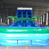 Front view of large inflatable water slide with tropical theme, featuring blue sliding lanes, palm tree decorations, and a colorful splash pool area with wave patterns. Set up in an indoor warehouse with metal roofing visible.