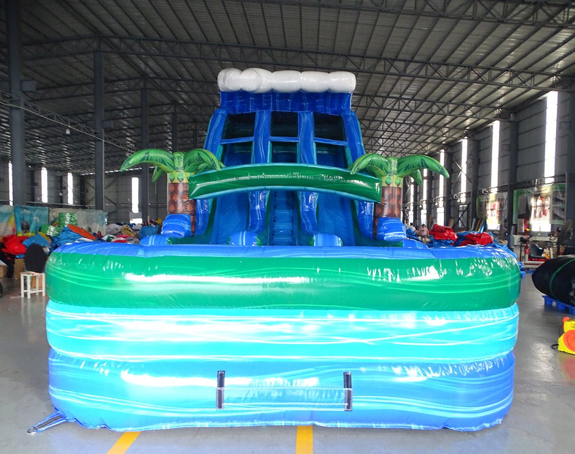 Front view of large inflatable water slide with tropical theme, featuring blue sliding lanes, palm tree decorations, and a colorful splash pool area with wave patterns. Set up in an indoor warehouse with metal roofing visible.