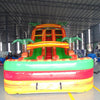 Large inflatable water slide with tropical theme, featuring orange and green colors, palm tree decorations, and three sliding lanes. Set up in an indoor warehouse with other inflatables and equipment visible in the background.