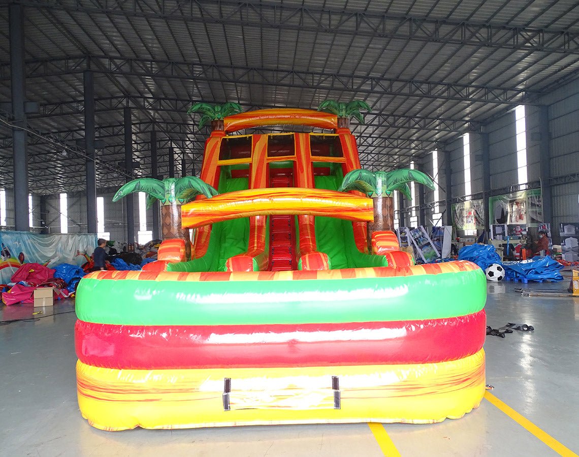 Large inflatable water slide with tropical theme, featuring orange and green colors, palm tree decorations, and three sliding lanes. Set up in an indoor warehouse with other inflatables and equipment visible in the background.