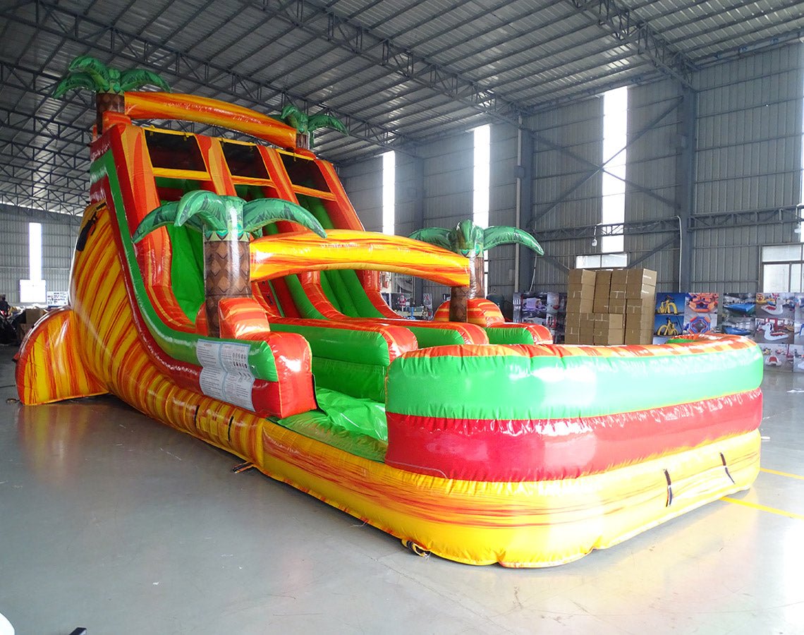 Side view of 18ft Tropical Inferno Center Climb water slide showing vibrant orange, green, and yellow colors, inflatable palm trees, and a large multi-colored splash pool area. The slide is displayed in an indoor facility with concrete flooring.