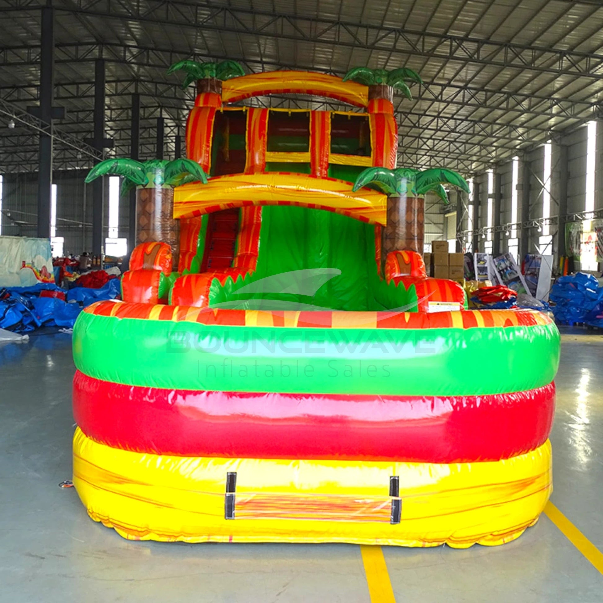 Front view of Tropical Inferno inflatable water slide displaying vibrant yellow, red, and green design, dual lanes, palm tree accents, and spacious splash pool with inflatable barriers.