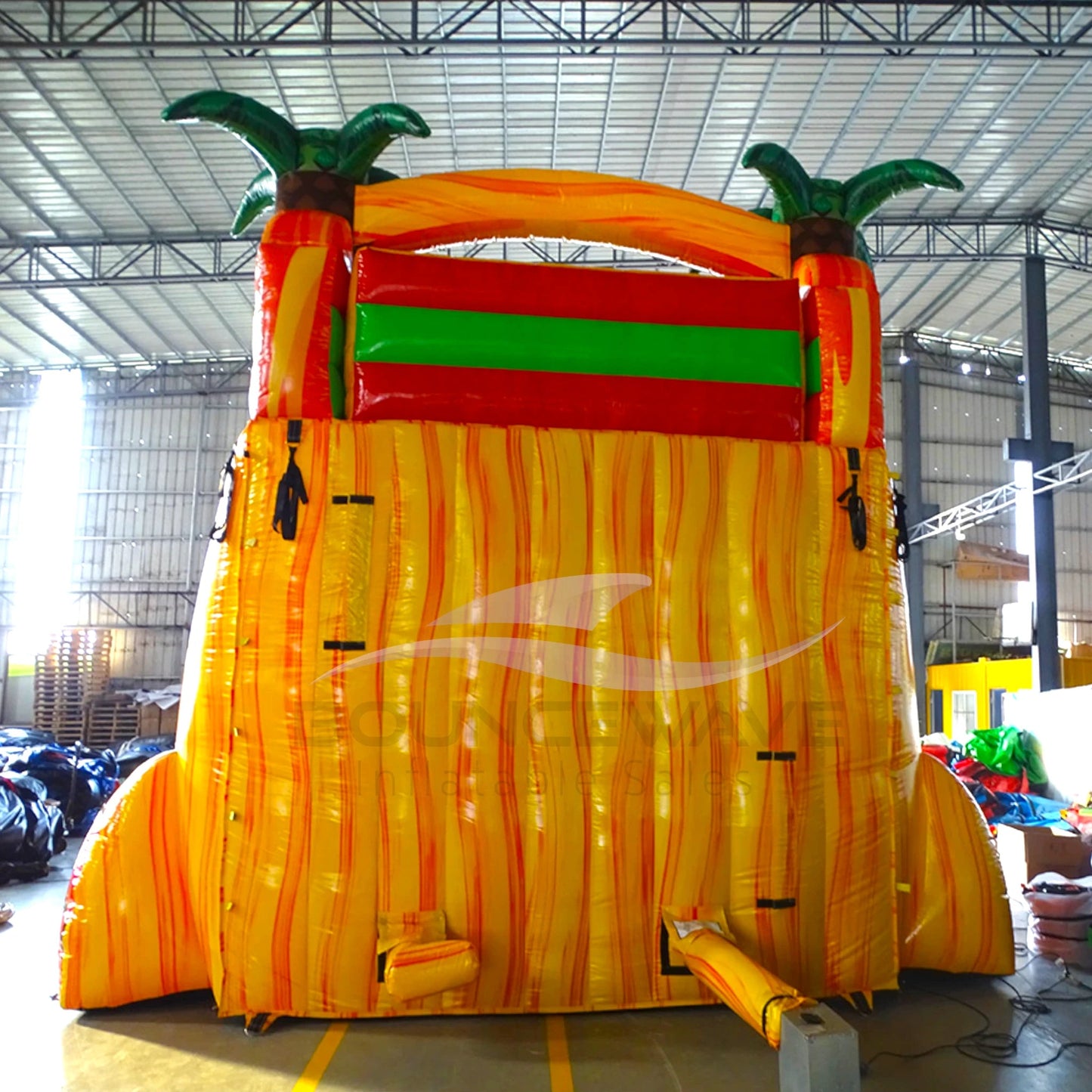 rear view Large tropical-themed inflatable water slide featuring palm tree decorations, dual yellow sliding lanes, and a vibrant orange and red color scheme in an indoor warehouse setting.