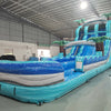 Side angle view of the 20' Bahama Blast Hybrid DL inflatable water slide, showcasing its blue sliding lanes, palm tree decorations, and colorful splash pool. The slide combines open and tube slide elements, set up in a spacious indoor warehouse with other equipment visible.