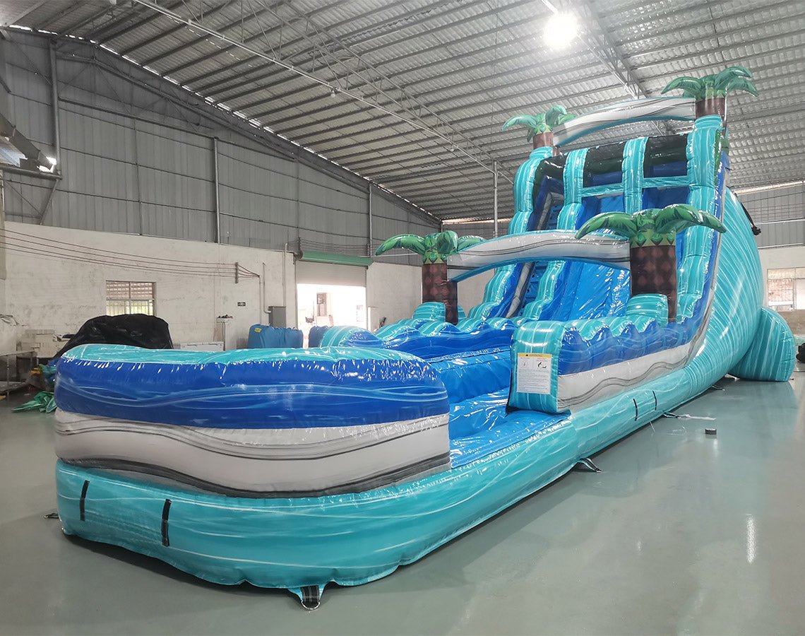 Side angle view of the 20' Bahama Blast Hybrid DL inflatable water slide, showcasing its blue sliding lanes, palm tree decorations, and colorful splash pool. The slide combines open and tube slide elements, set up in a spacious indoor warehouse with other equipment visible.