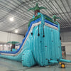 Rear view of 20' Bahama Blast Hybrid DL water slide, displaying its blue and turquoise color scheme, inflatable palm trees, and enclosed tube slide section. Set up in an indoor warehouse with metal roofing and other inflatables visible in the background.