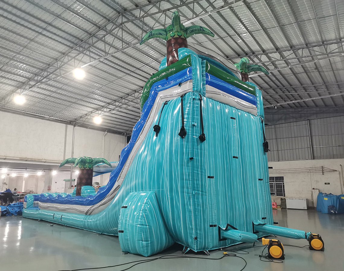 Rear view of 20' Bahama Blast Hybrid DL water slide, displaying its blue and turquoise color scheme, inflatable palm trees, and enclosed tube slide section. Set up in an indoor warehouse with metal roofing and other inflatables visible in the background.